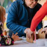 two person making gingerbread house and three children watching them